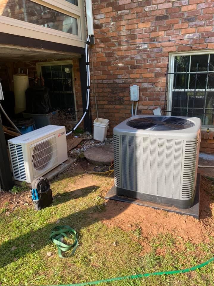 Two air conditioning units sit on the ground outside a brick house, one rectangular and horizontal, the other square.