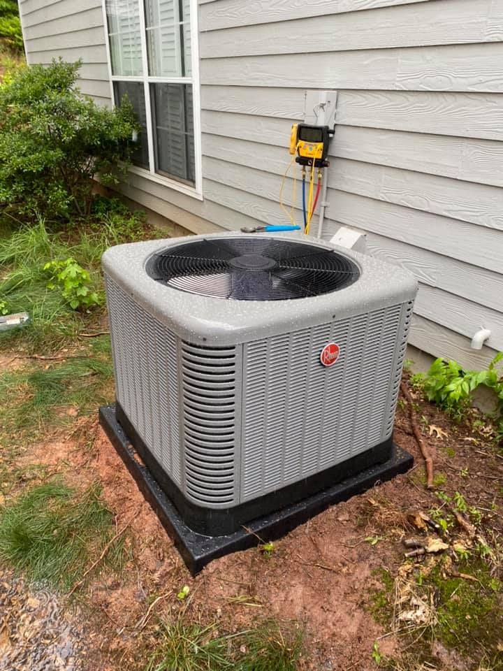 A grey Rheem air conditioning condenser unit sits on a black pad outside a house, with diagnostic gauges attached to it.