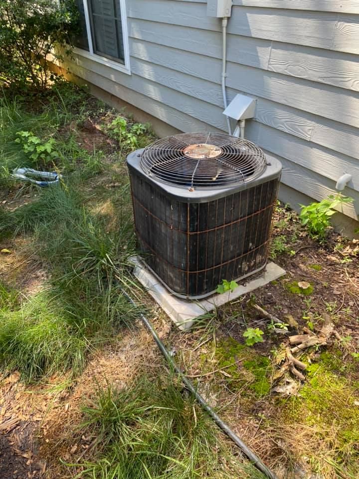 An air conditioning condenser unit sits on a concrete pad next to a house with gray siding surrounded by dirt and weeds.