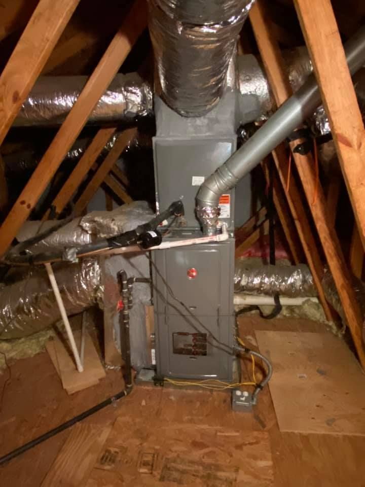 An HVAC air handler unit installed in an attic with insulated ductwork, metal flue pipe, and wooden rafters.
