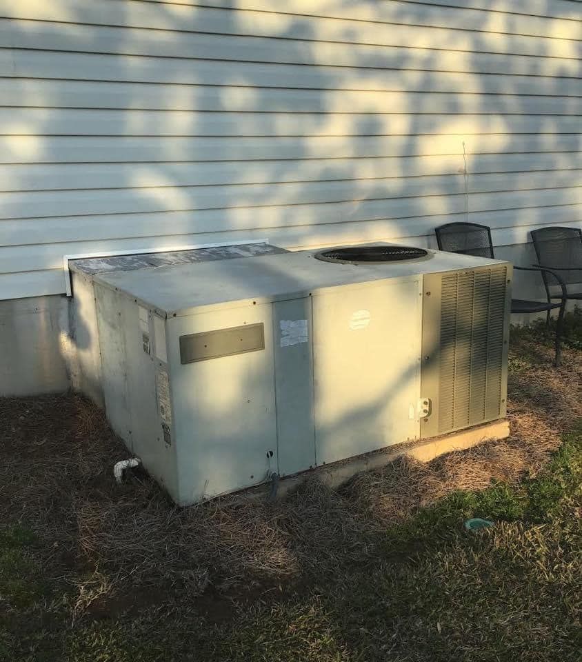 A light-colored, rectangular HVAC unit sits outdoors against a white siding wall with tree-shadow patterns.