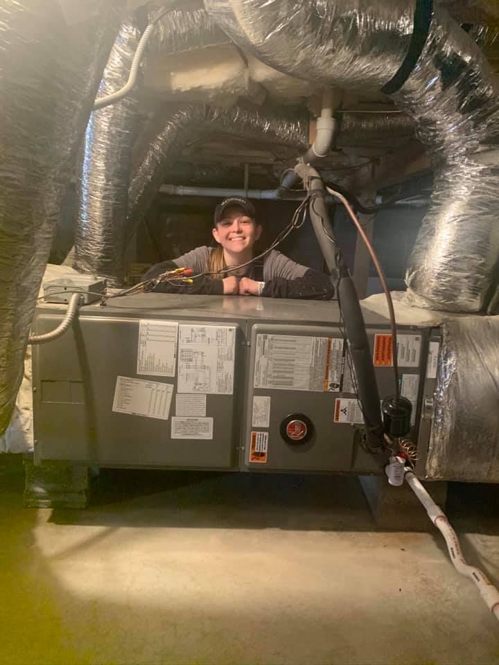A person wearing a baseball cap smiles while leaning against a large HVAC unit in a crawl space.