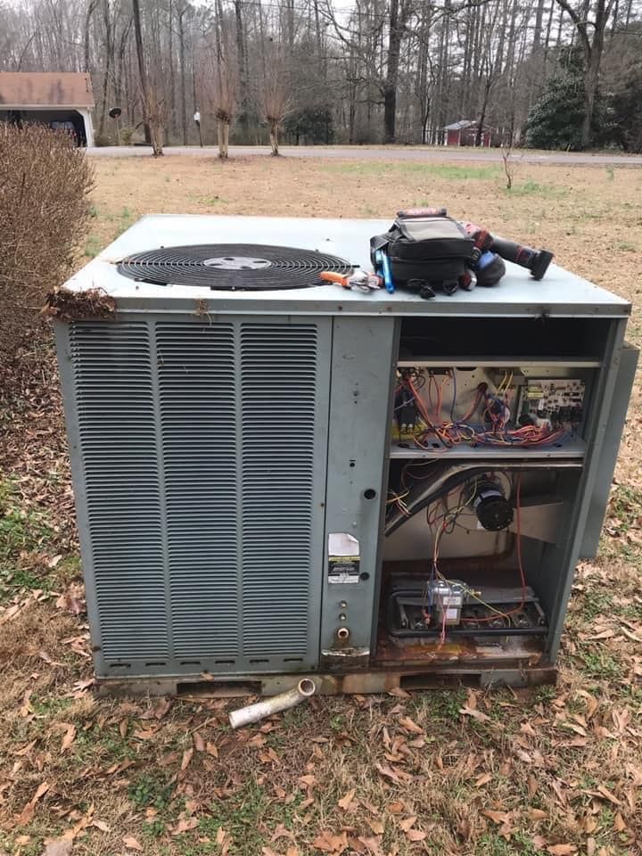A grey HVAC unit sits outdoors on a lawn with its side panel open, exposing internal wiring and components.