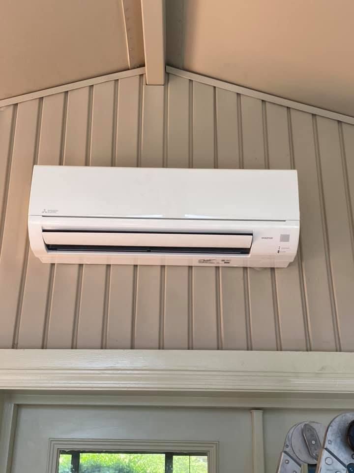 A white wall-mounted air conditioner unit installed on a beige vertical-paneled wall above a window and door frame.