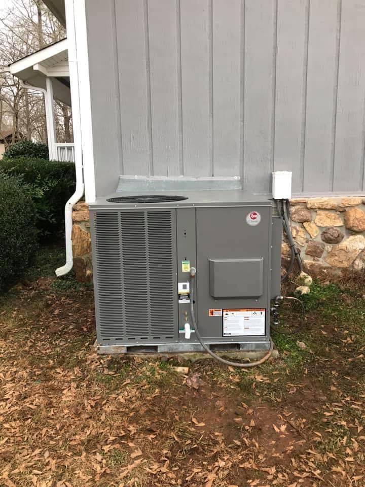 A gray outdoor HVAC unit sits on a small concrete pad next to the stone foundation and gray siding of a house.