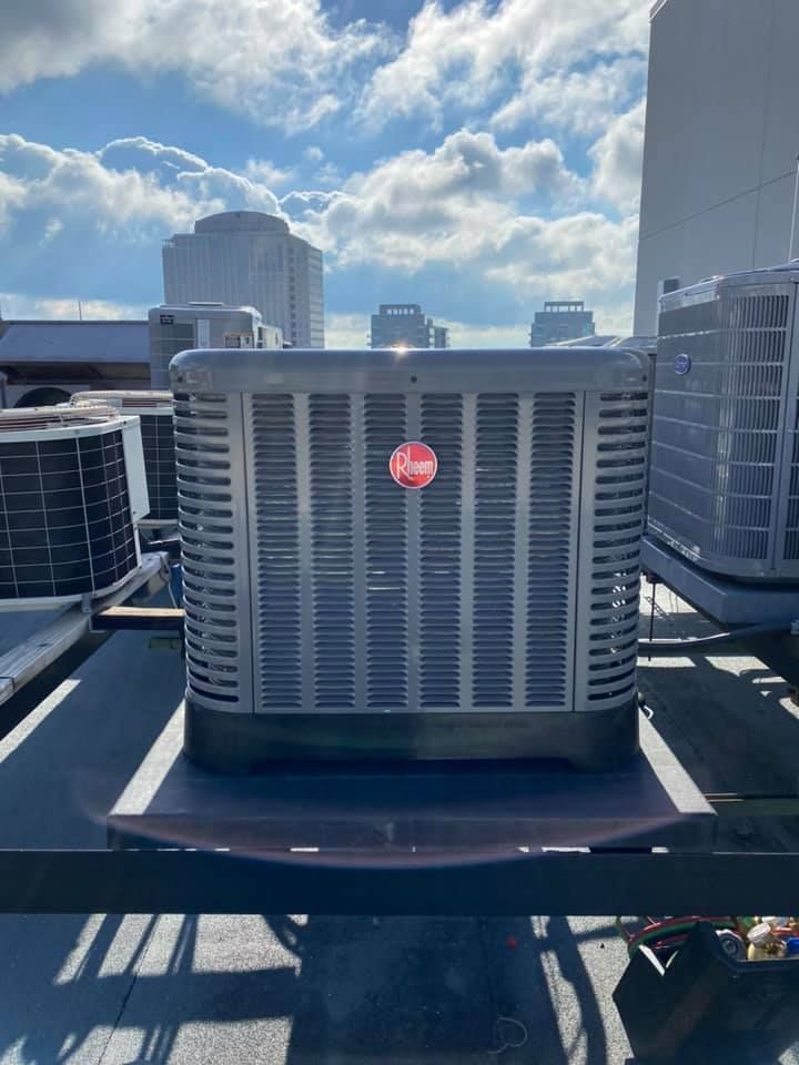 A grey Rheem outdoor HVAC condenser unit on a rooftop, set against a bright, cloudy sky with distant city buildings.