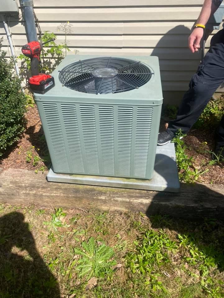 A technician standing next to a light green central air conditioning unit outdoors, with a red power tool resting on top.