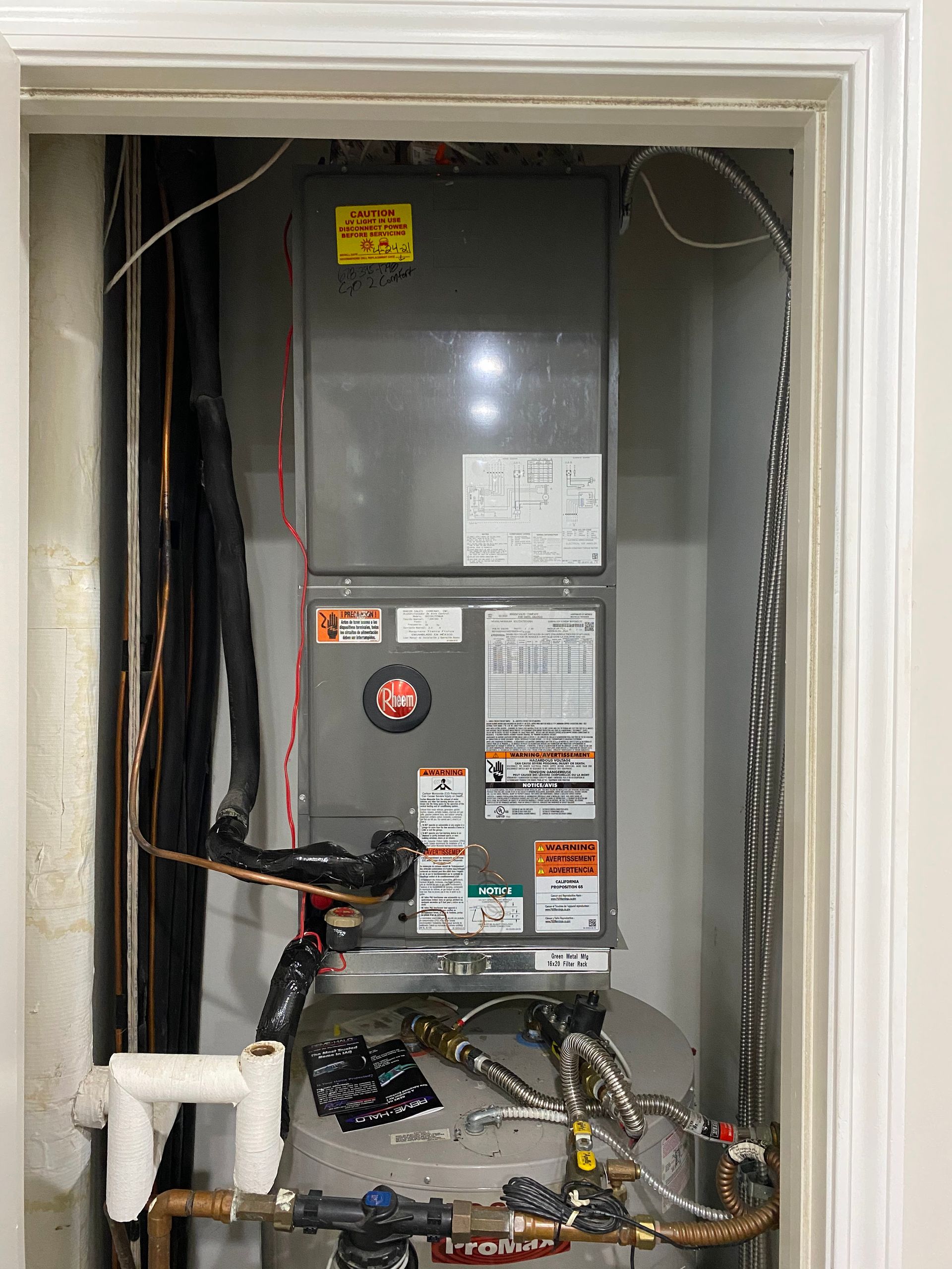 A central HVAC air handler unit installed in a closet above a water heater, with various pipes and electrical wiring.