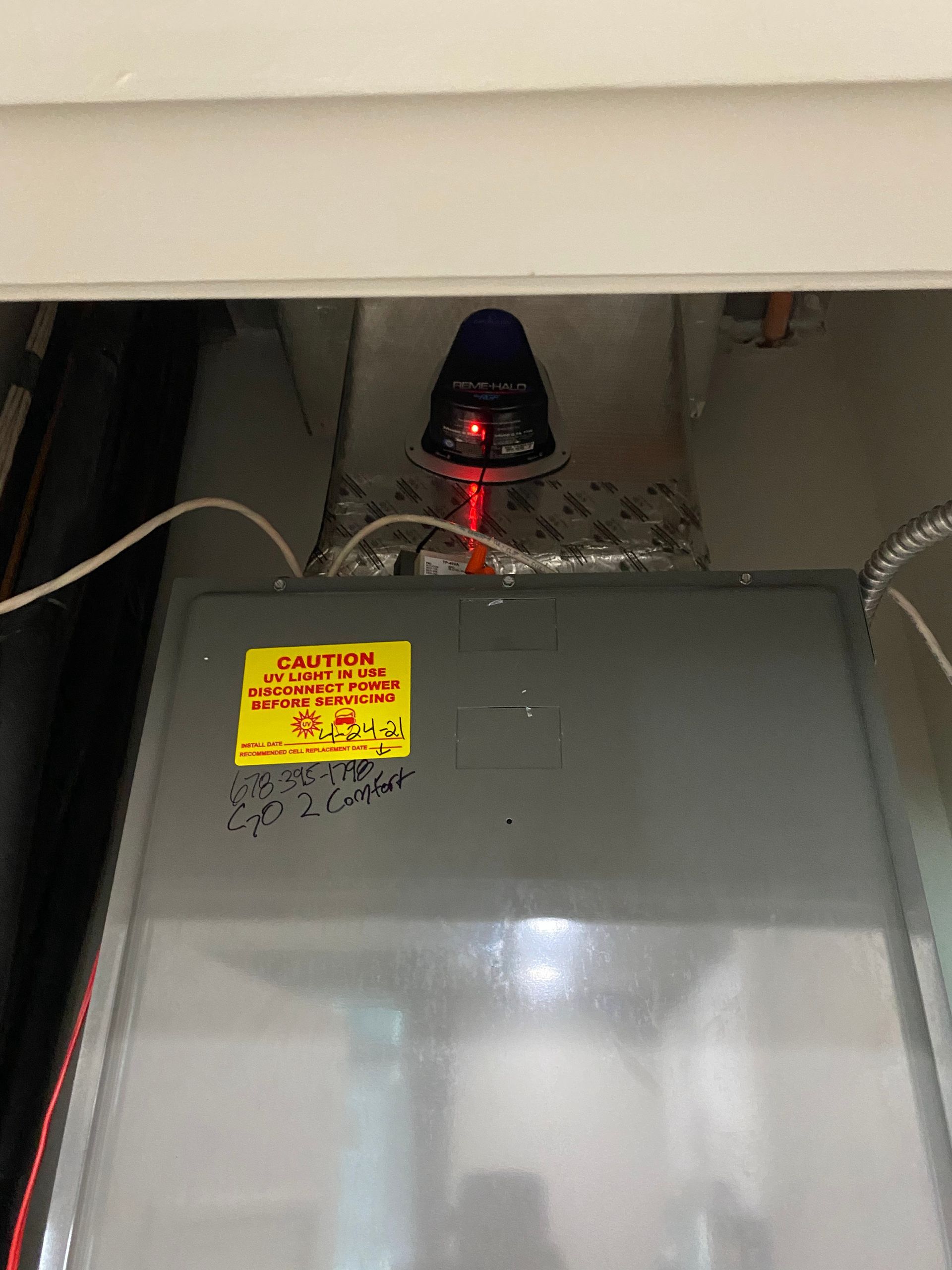 A residential HVAC duct-mounted smoke detector with a glowing red LED light mounted on metal ductwork above a furnace.