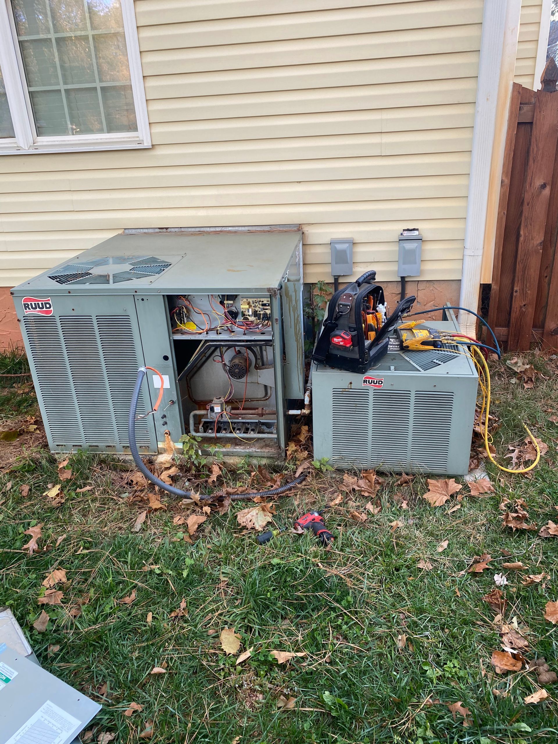 Two HVAC units sitting on the grass outside a house, with a technician's tool bag resting on top of one unit.