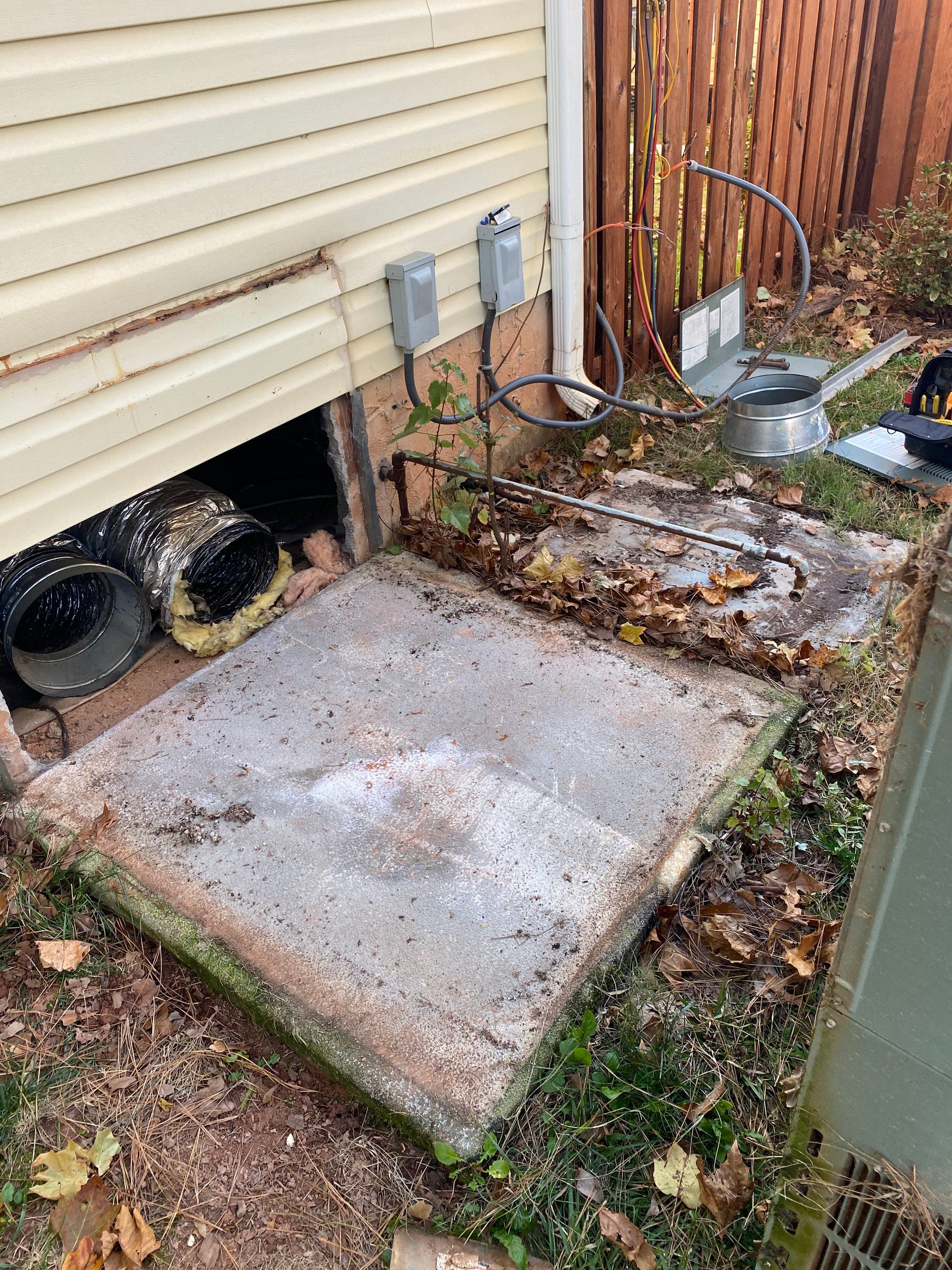 A concrete pad beside a house exterior with an open crawlspace access, electrical conduit, and disconnected ductwork.