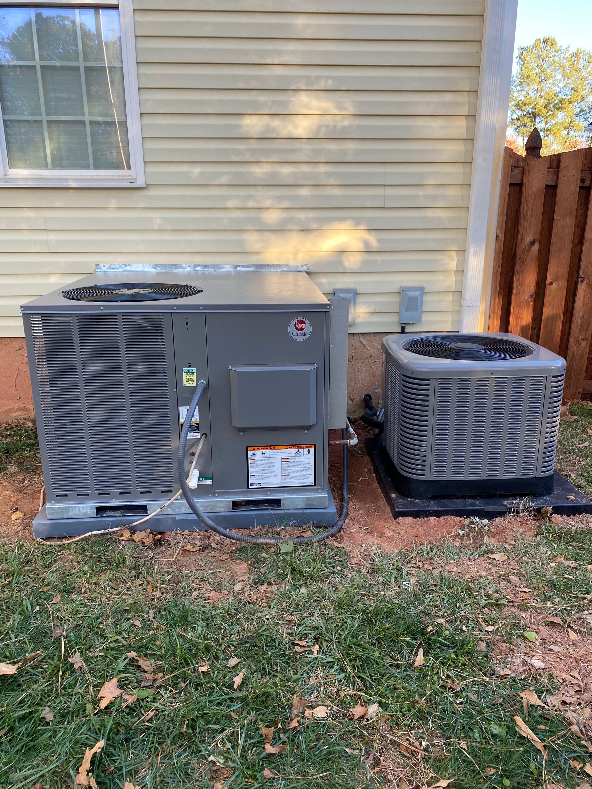 Two HVAC units sit on a dirt patch beside a house with light-colored siding and a wooden fence in the background.