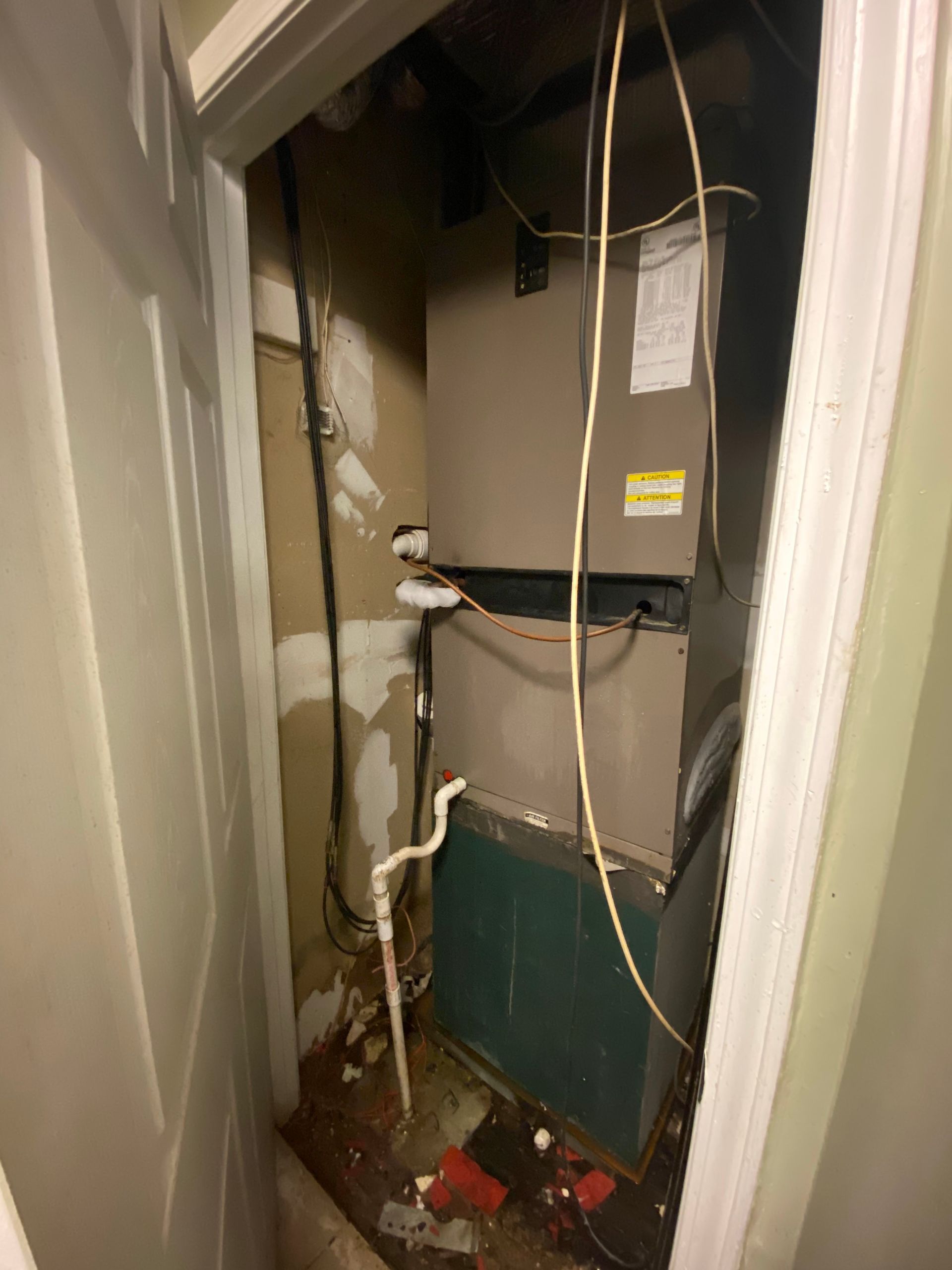 A vertical HVAC unit in a small, unfinished closet with exposed wiring and PVC piping.