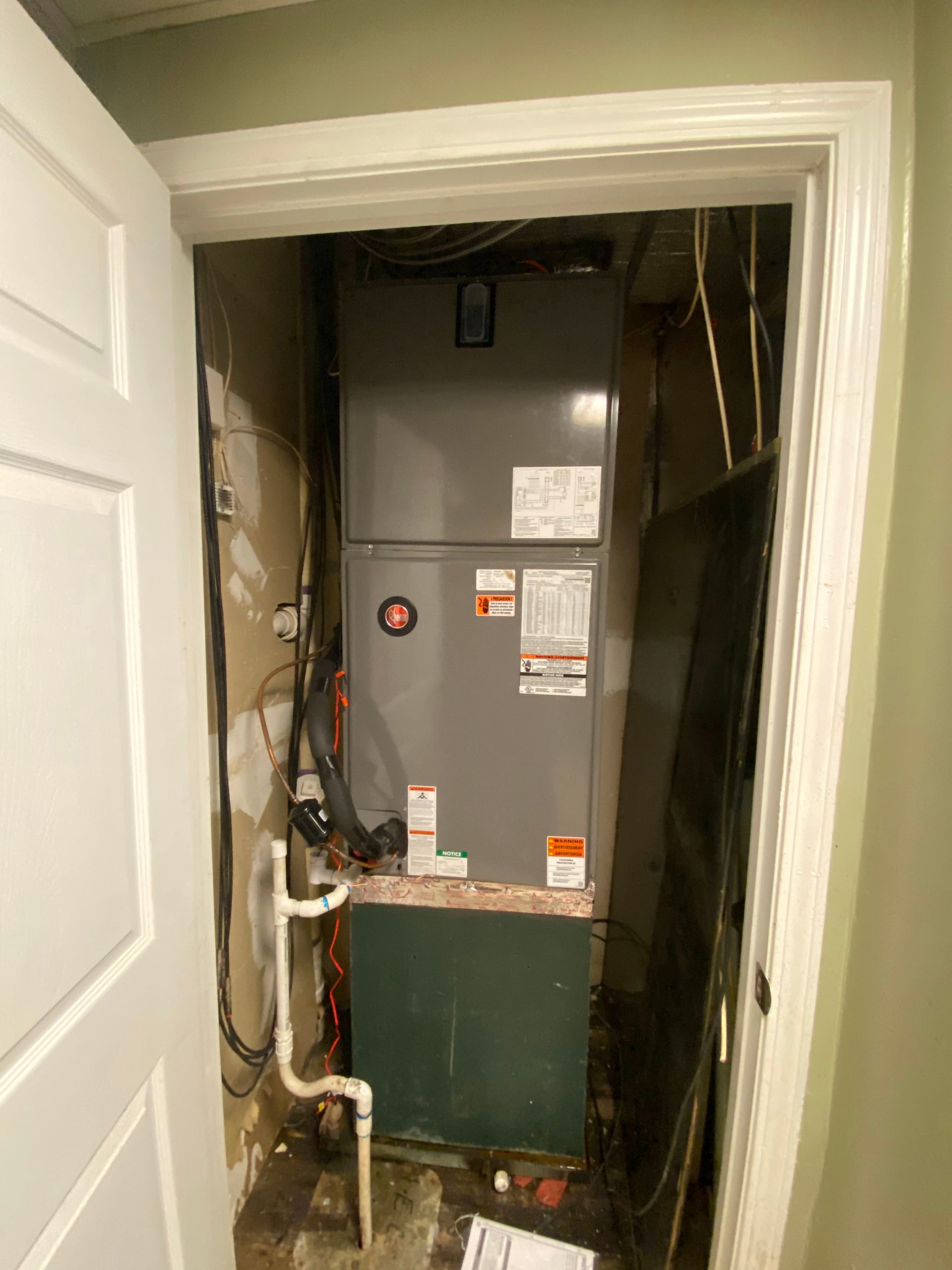 A gray HVAC air handler unit installed in a utility closet with exposed wiring and white drainage piping.