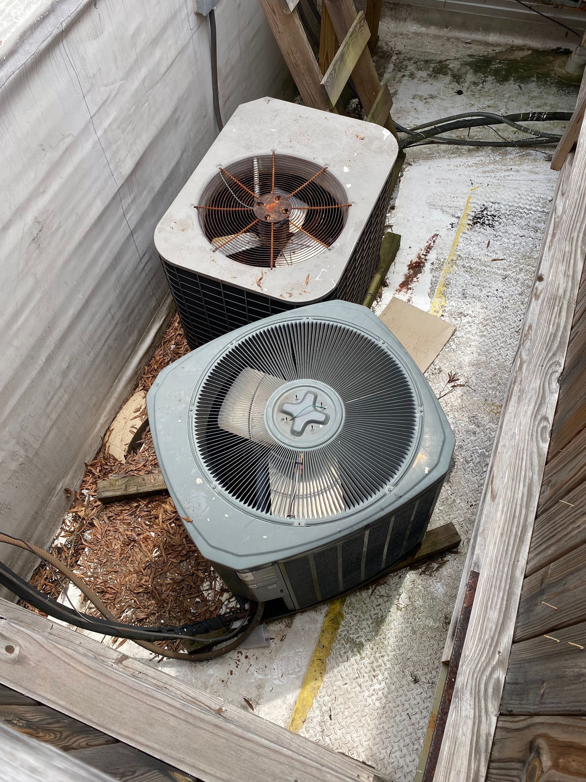 Two outdoor air conditioning condenser units sit side-by-side on a concrete pad enclosed by wooden walls.