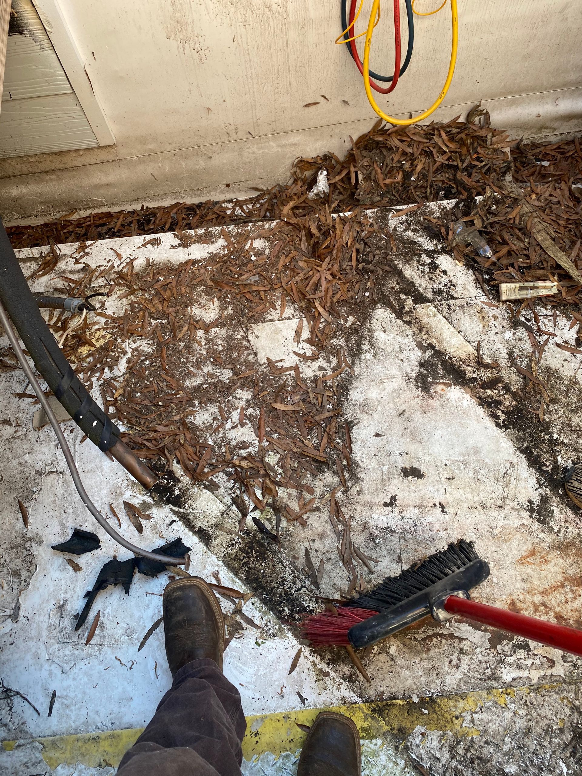A person uses a broom to clear dirt and debris from a workshop floor, with a yellow cord hanging in the background.