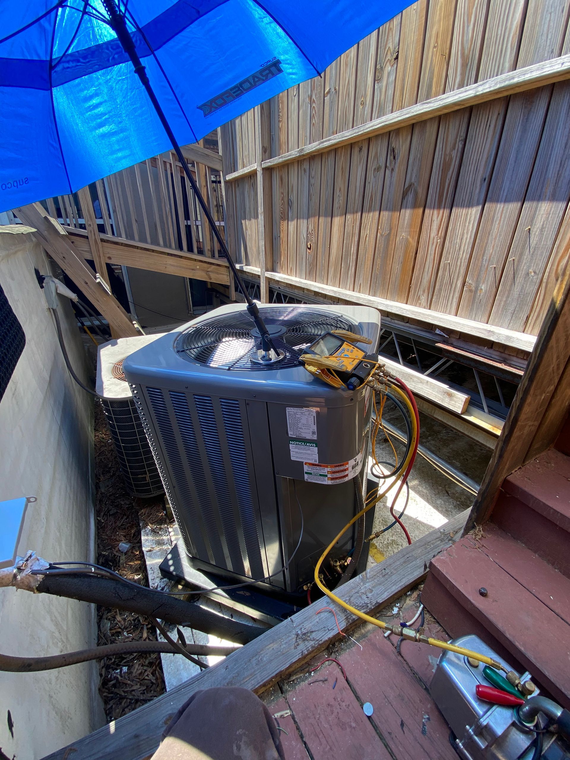 An HVAC technician works on a grey outdoor AC unit on a wooden deck, using gauges and a vacuum pump under a blue umbrella.