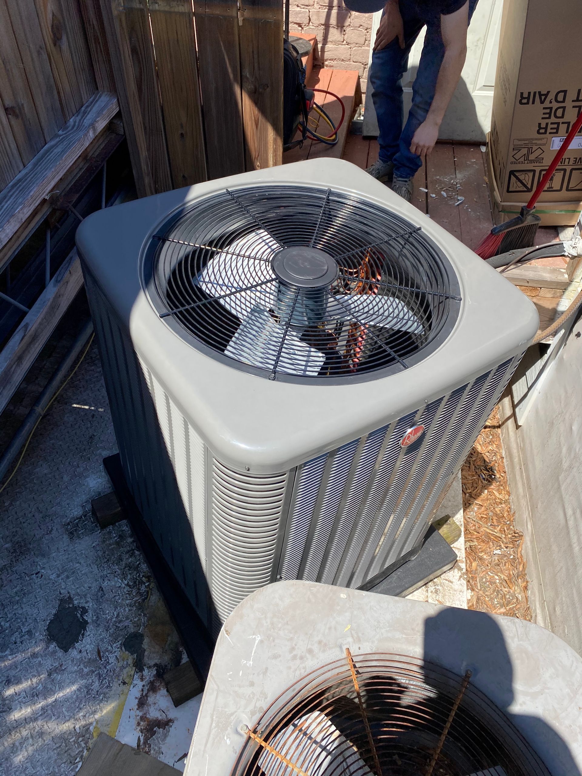 An outdoor air conditioning unit with a fan visible through a top grille, with a partial view of a person standing nearby.