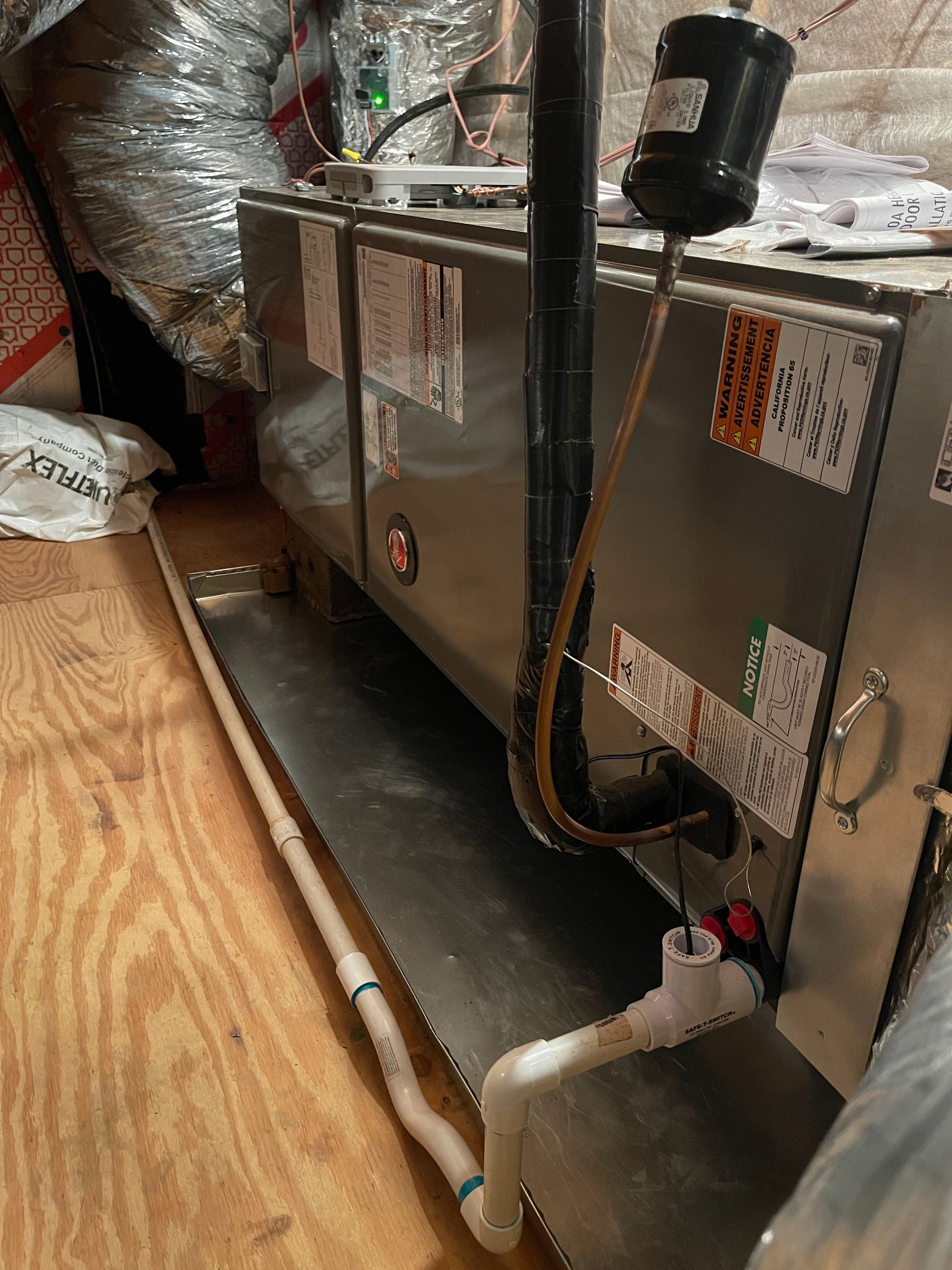 A metallic HVAC air handler unit installed in an attic, featuring connected white PVC drain pipes and copper tubing.