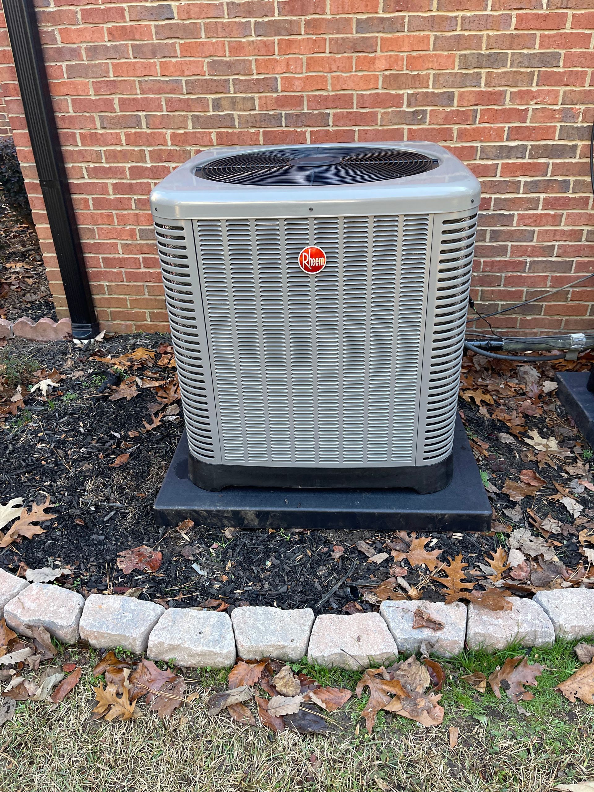 A Rheem central air conditioning unit sits on a black pad in a mulched garden bed against a red brick wall.