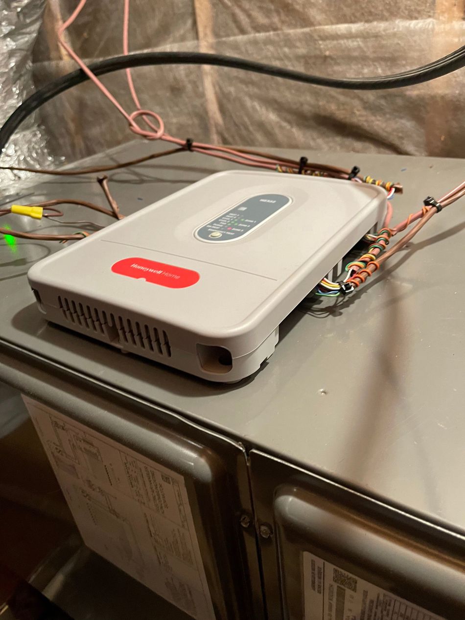 A light gray Aprilaire air purifier controller mounted on top of a gray furnace unit with exposed wiring.