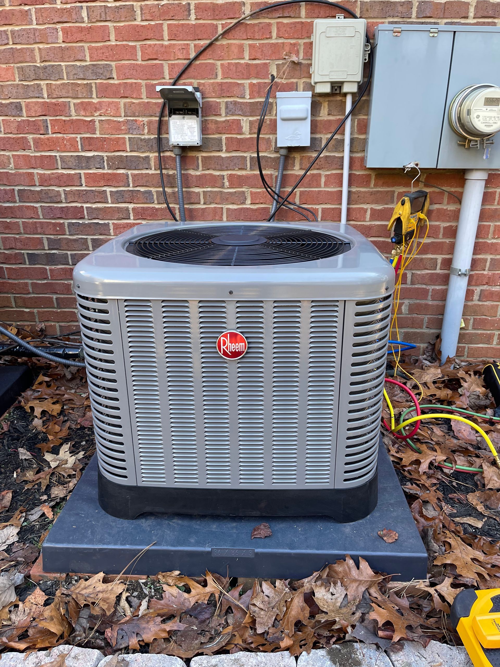 A gray Rheem outdoor air conditioning unit sits on a dark plastic pad against a brick wall with electrical conduits.