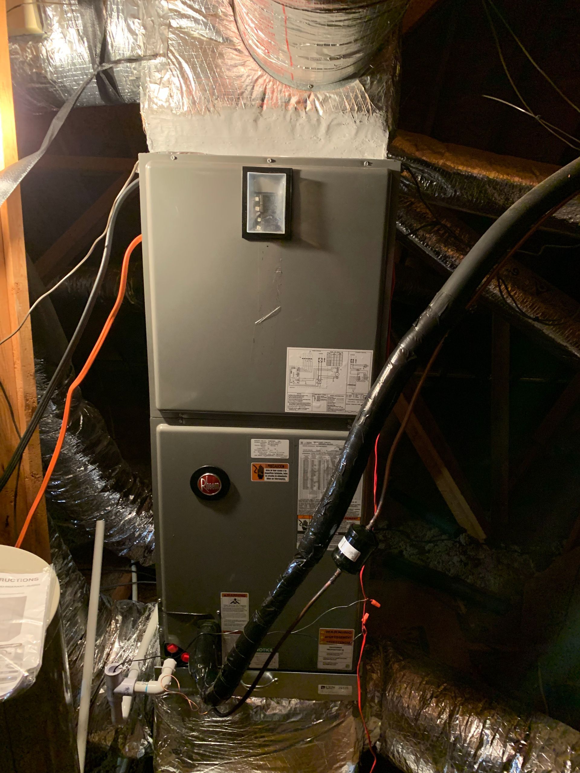 An HVAC air handler unit installed in an attic space, with connected silver ductwork and various electrical wires.