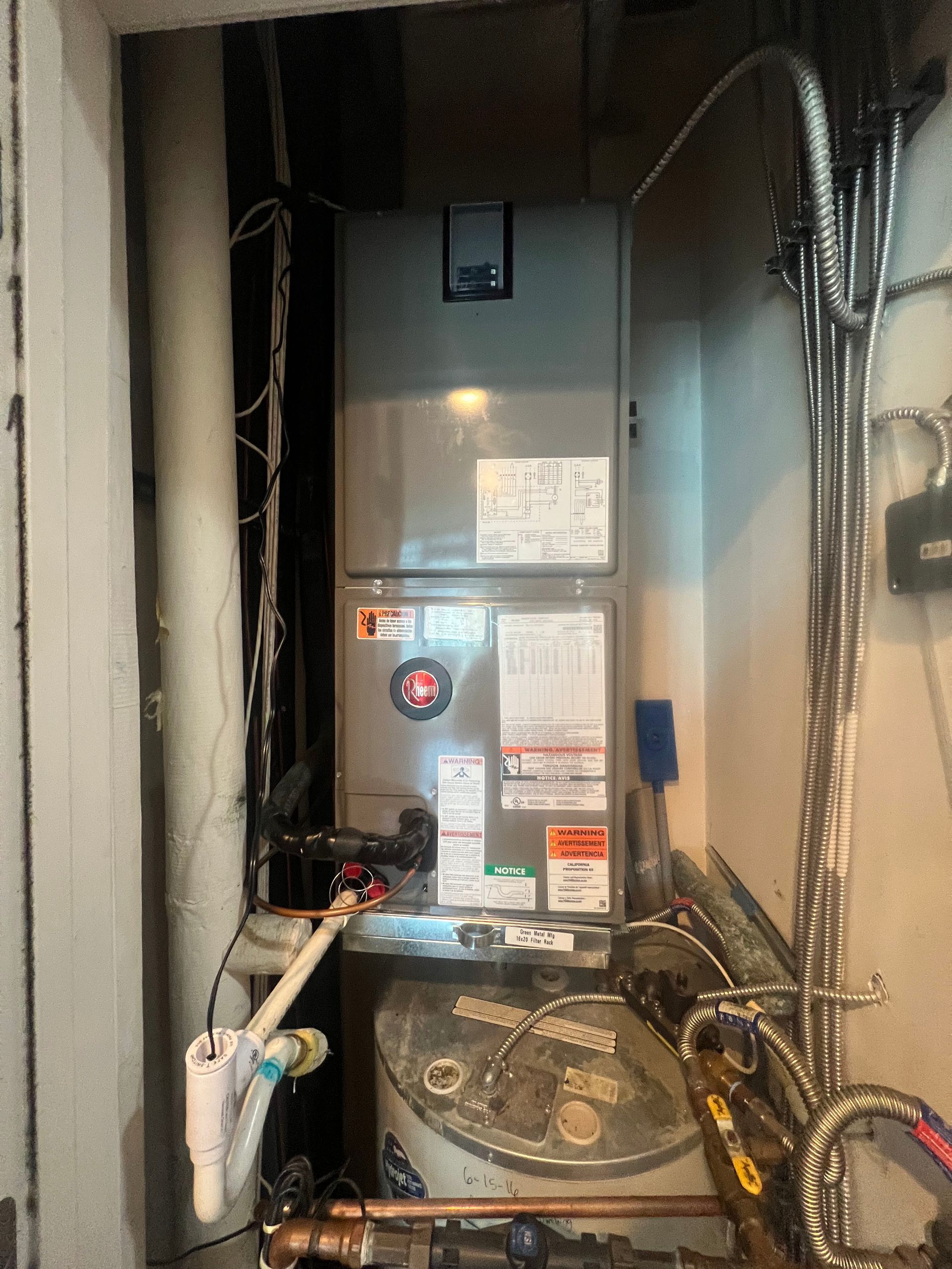 A grey HVAC air handler unit installed in a utility closet, positioned above a water heater with visible pipe connections.