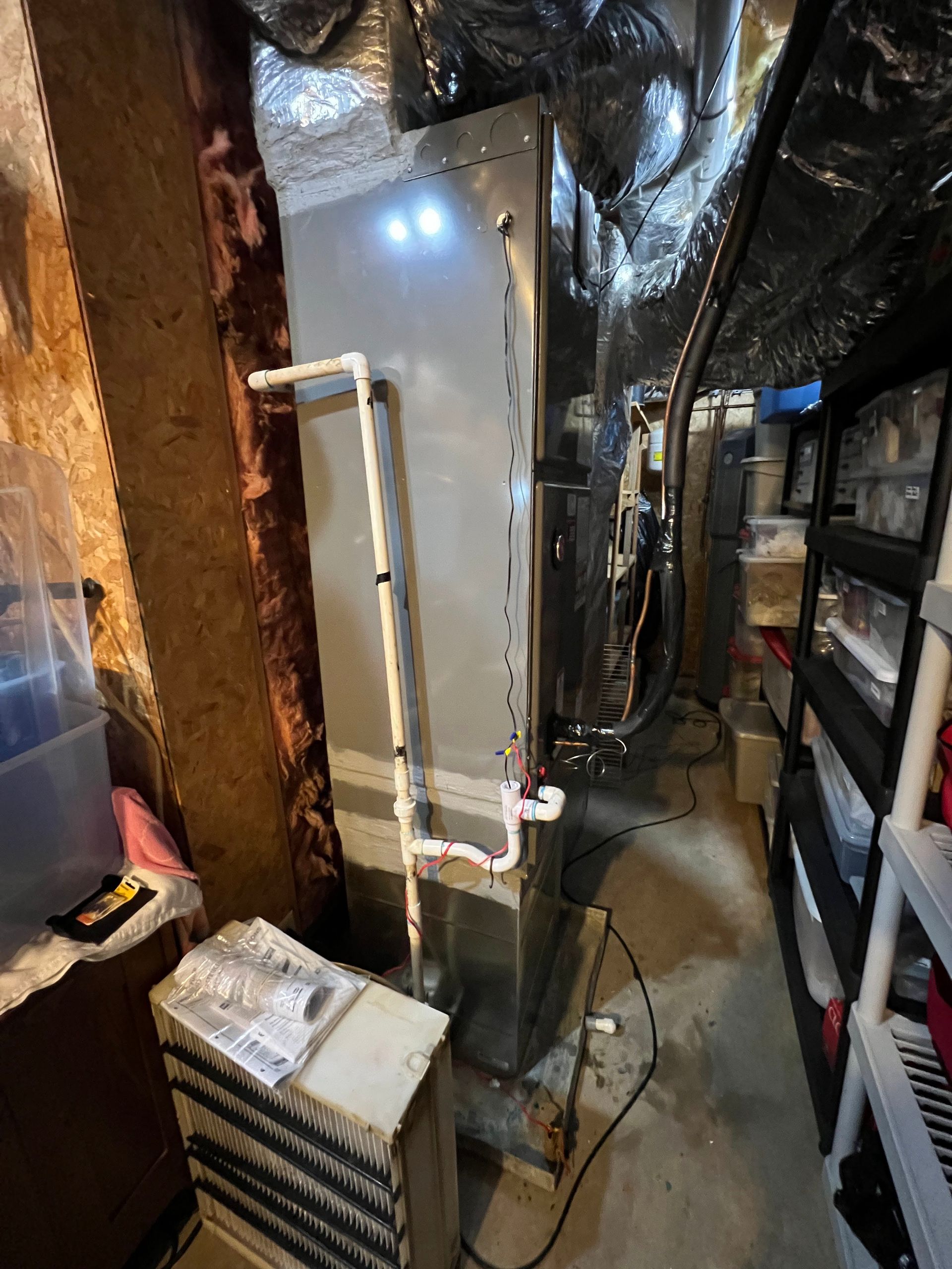 A vertical gray furnace unit in a basement with white PVC drain pipes attached to its side, near shelving and ductwork.