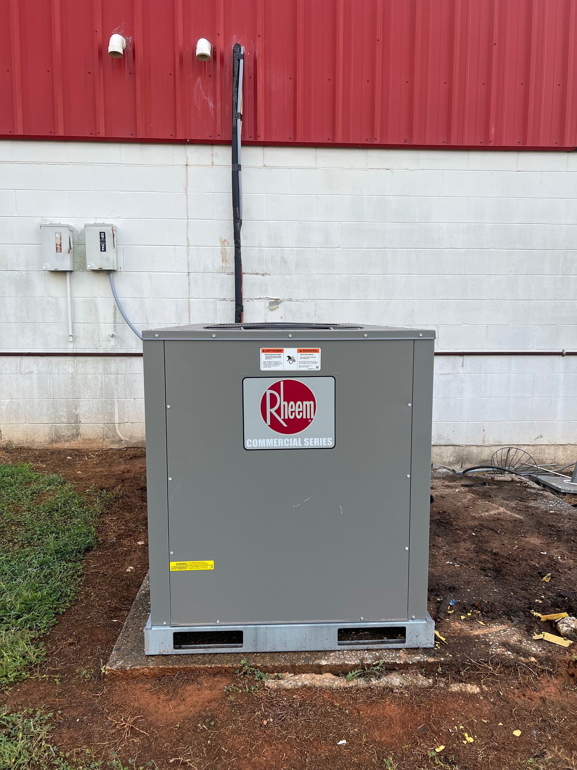 A grey Rheem HVAC unit sits on a concrete pad against a white cinder block wall under a red metal building exterior.