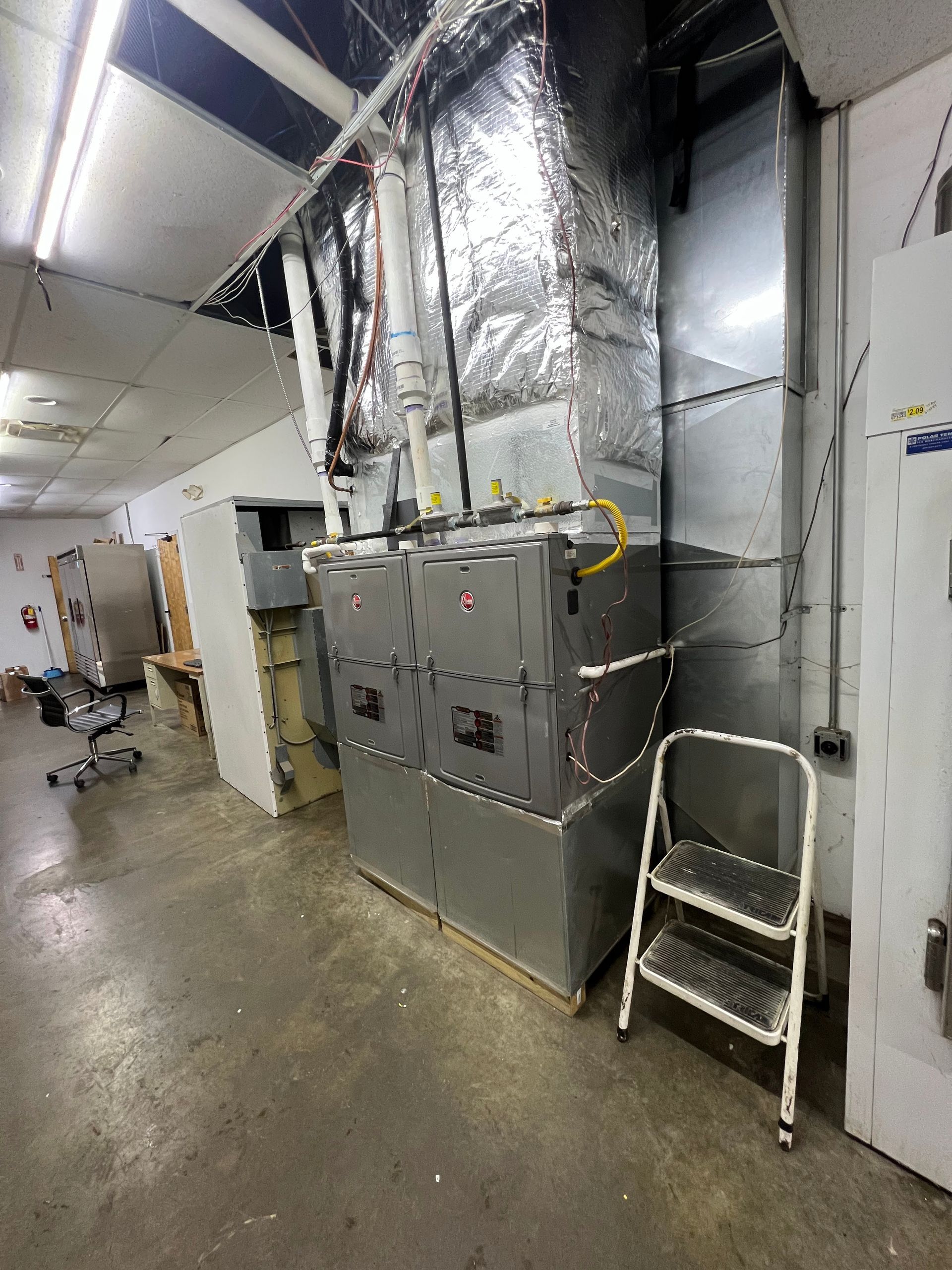 A large, metal HVAC unit stands in a utility room, featuring insulated ductwork and a small step ladder positioned nearby.