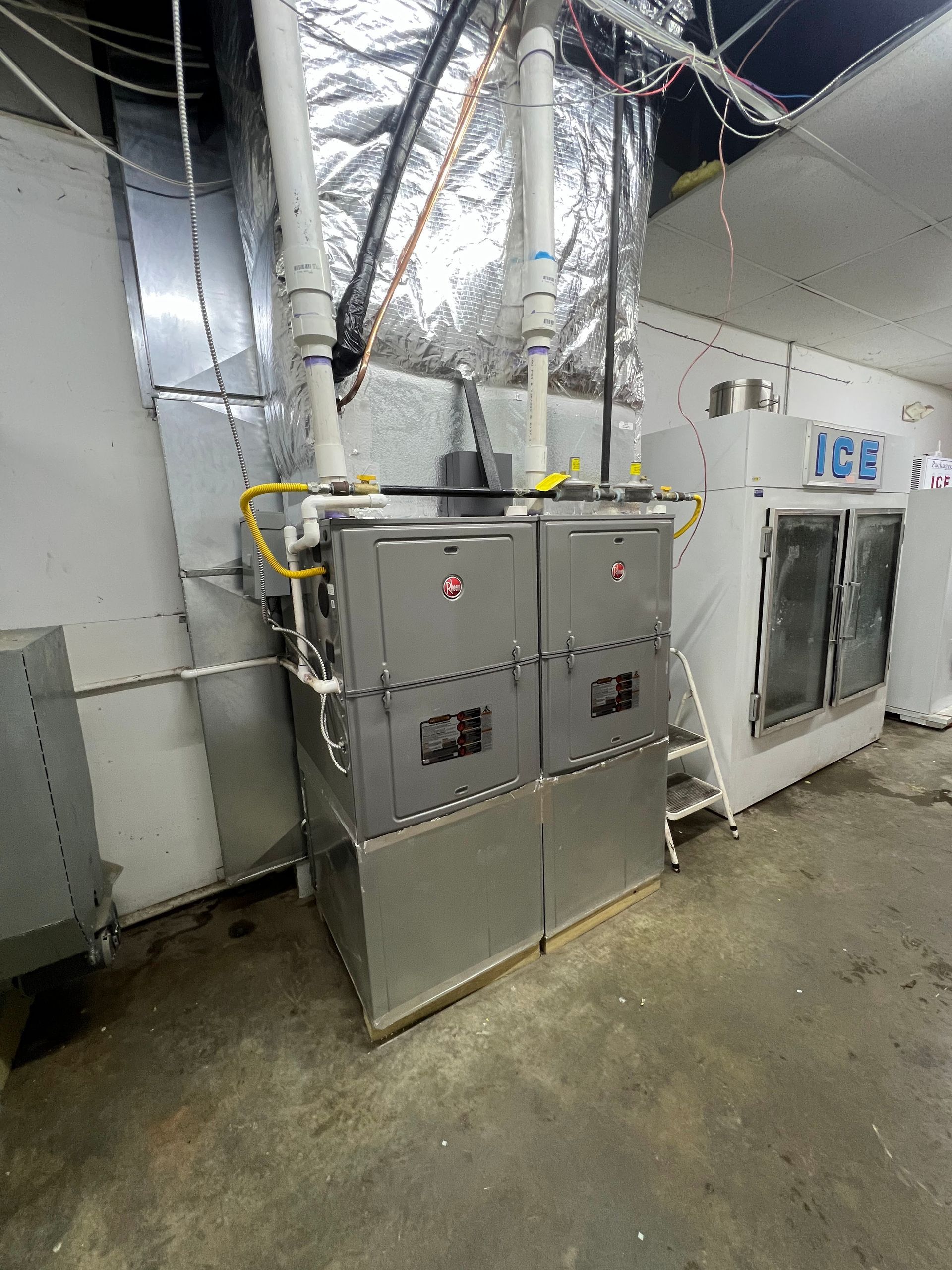 Two grey HVAC furnace units stand side-by-side in an industrial room, connected to white exhaust pipes and metal ductwork.