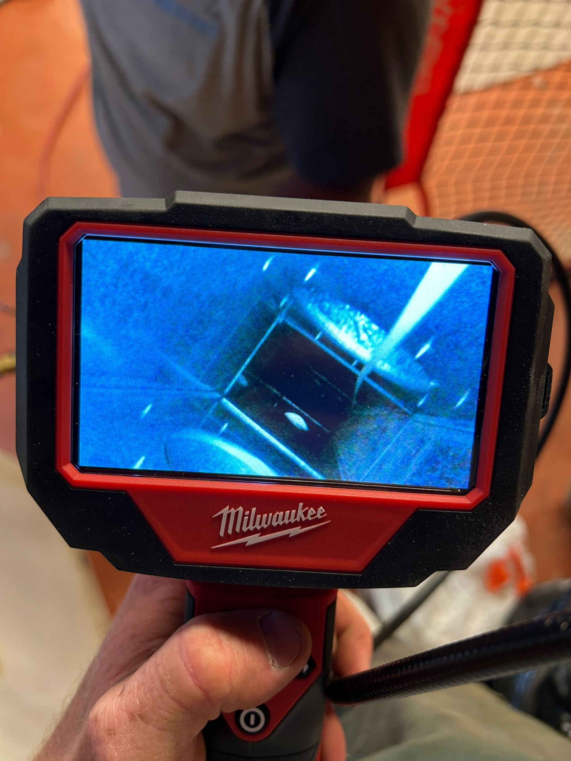 A person holds a Milwaukee inspection camera, showing a view of the inside of a dark, narrow pipe or drain on its screen.