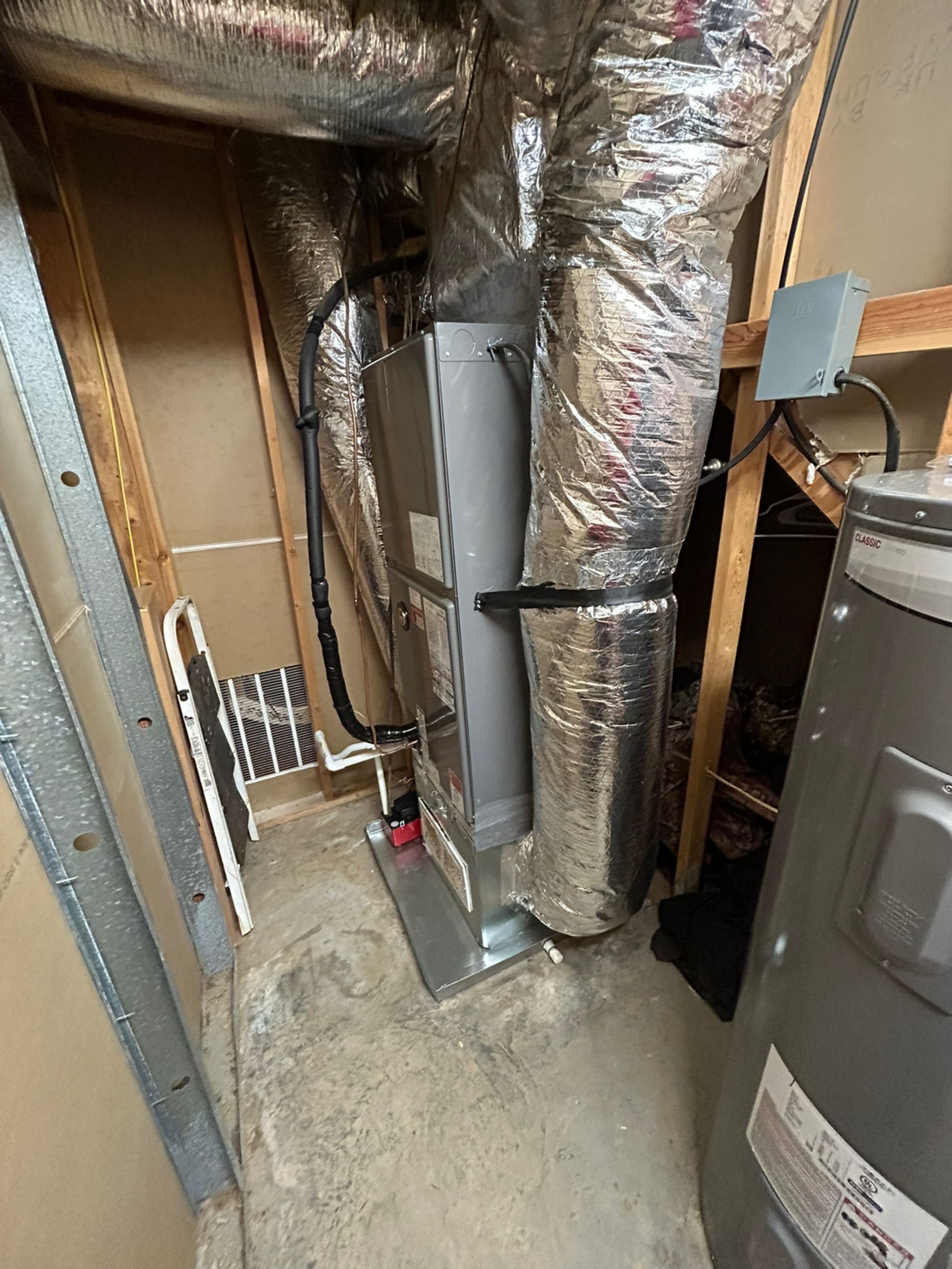 A vertical HVAC air handler unit with silver ductwork stands in a utility closet next to a water heater.