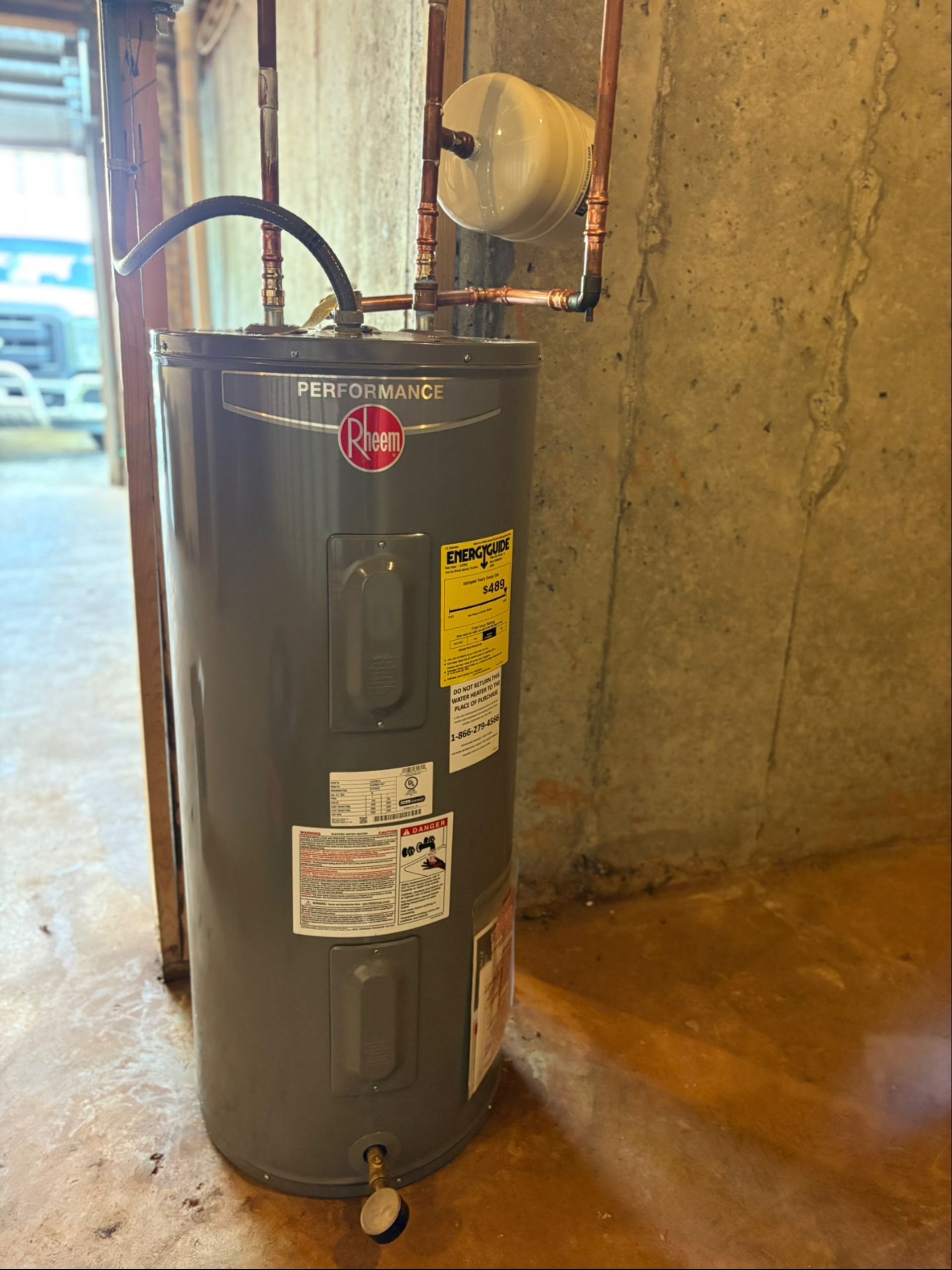 A gray Rheem electric water heater with copper plumbing connections, standing against a concrete wall in a garage.