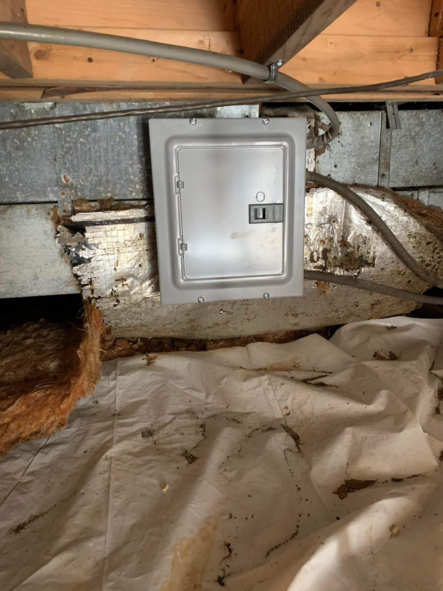 A gray electrical breaker box mounted on a foundation wall in a crawl space with a white vapor barrier on the ground.