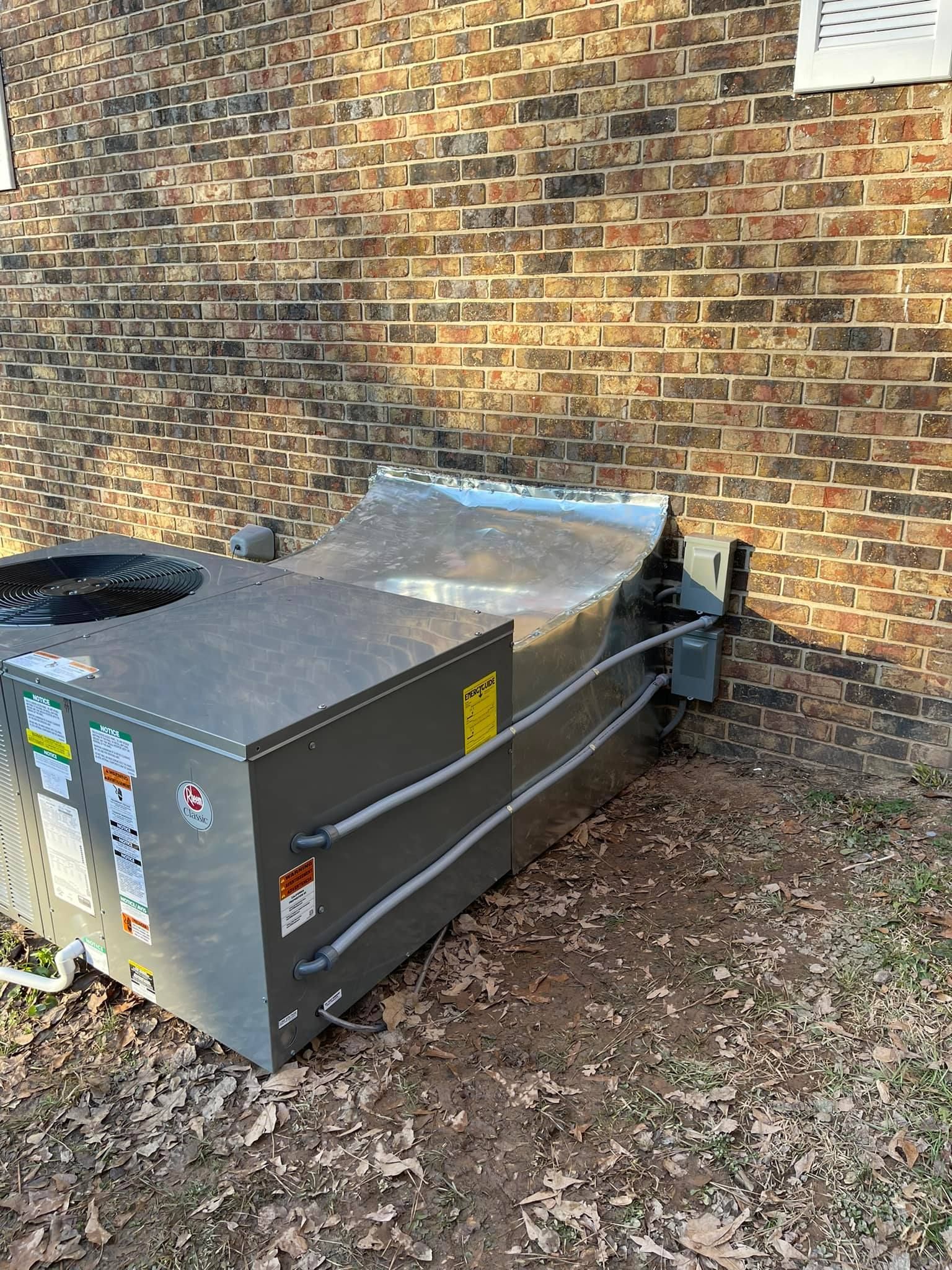A grey, rectangular HVAC unit sits on the ground next to a brown brick wall, connected by metal conduits.