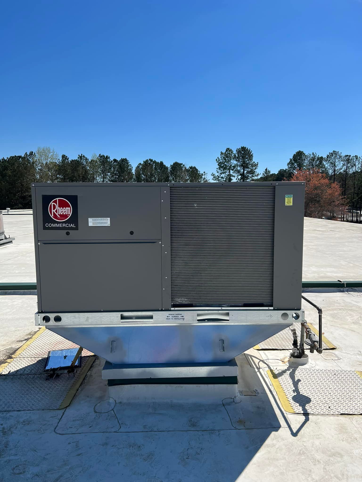 A grey commercial rooftop HVAC unit sits on a metal mounting frame under a clear blue sky.