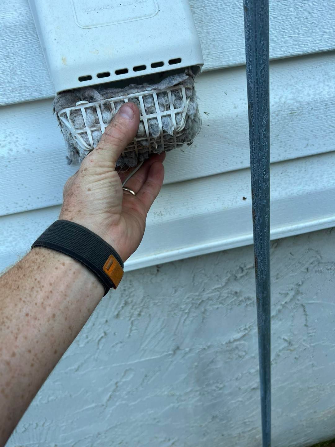 A hand pulls a white, plastic mesh guard clogged with gray lint away from a home exterior dryer vent.
