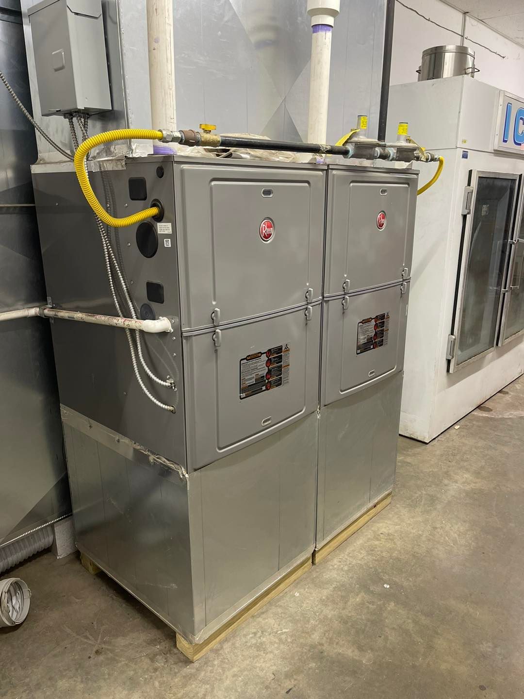 Two side-by-side stainless steel commercial furnace units with yellow gas lines, connected by chains in a workshop.