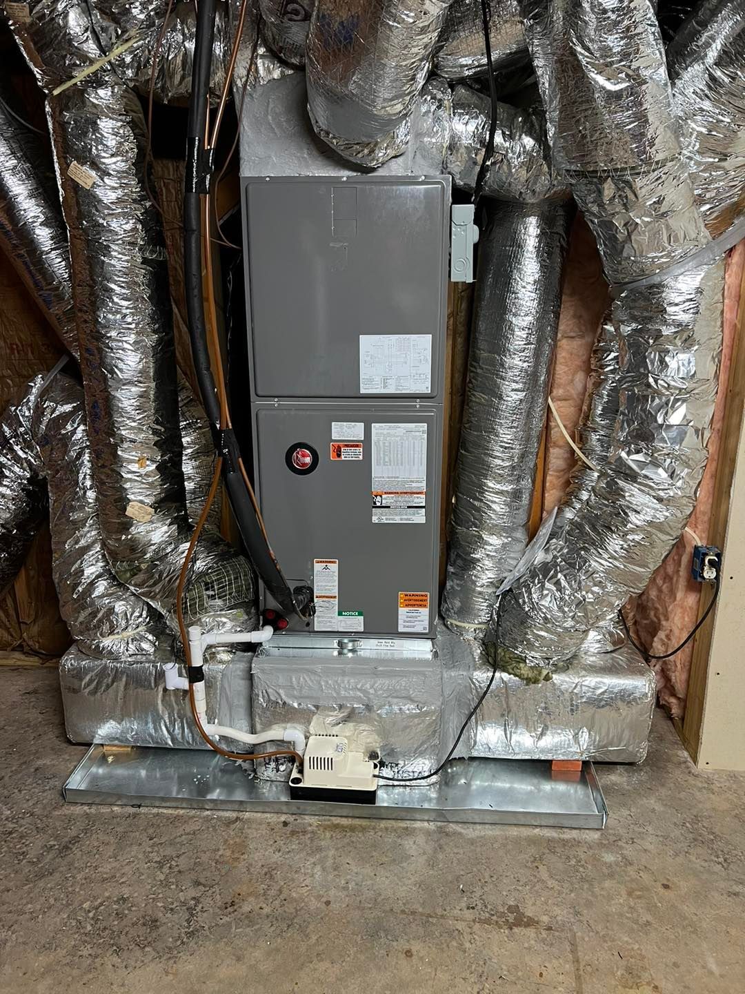 A gray HVAC air handler unit in a utility space, surrounded by silver insulated flexible ducts and a condensate pump.