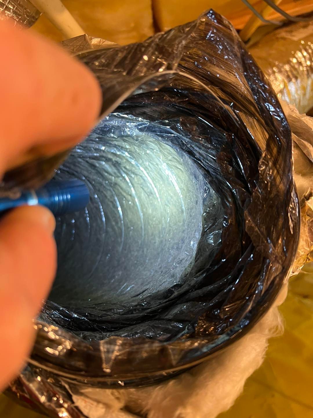 A hand shines a flashlight into the dark interior of a flexible, ribbed HVAC ducting tube.
