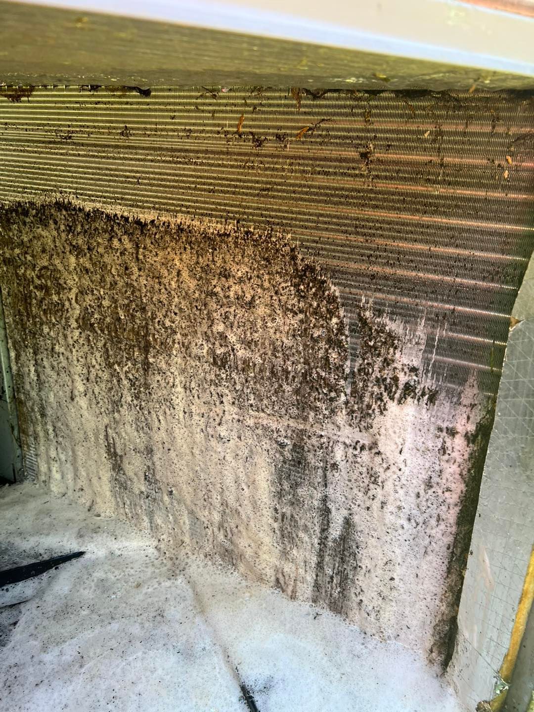 Dirty air conditioner condenser fins covered in thick, dark debris with white cleaning foam applied at the bottom.