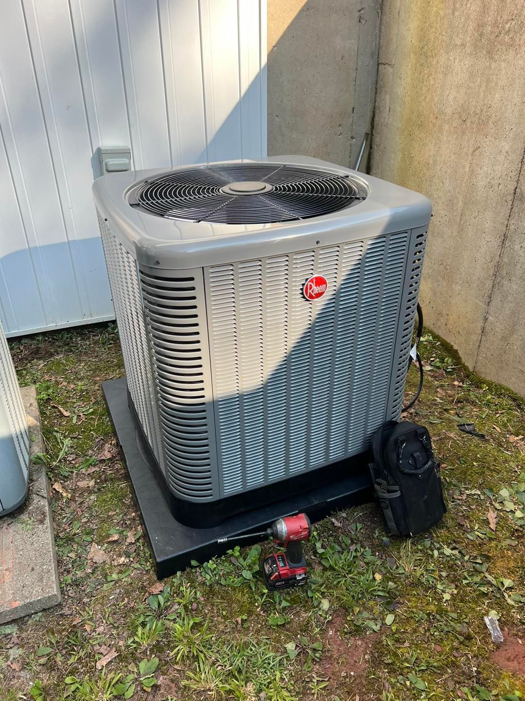 A Rheem outdoor air conditioning unit sits on a black pad in a yard next to a power drill and a disconnected panel cover.