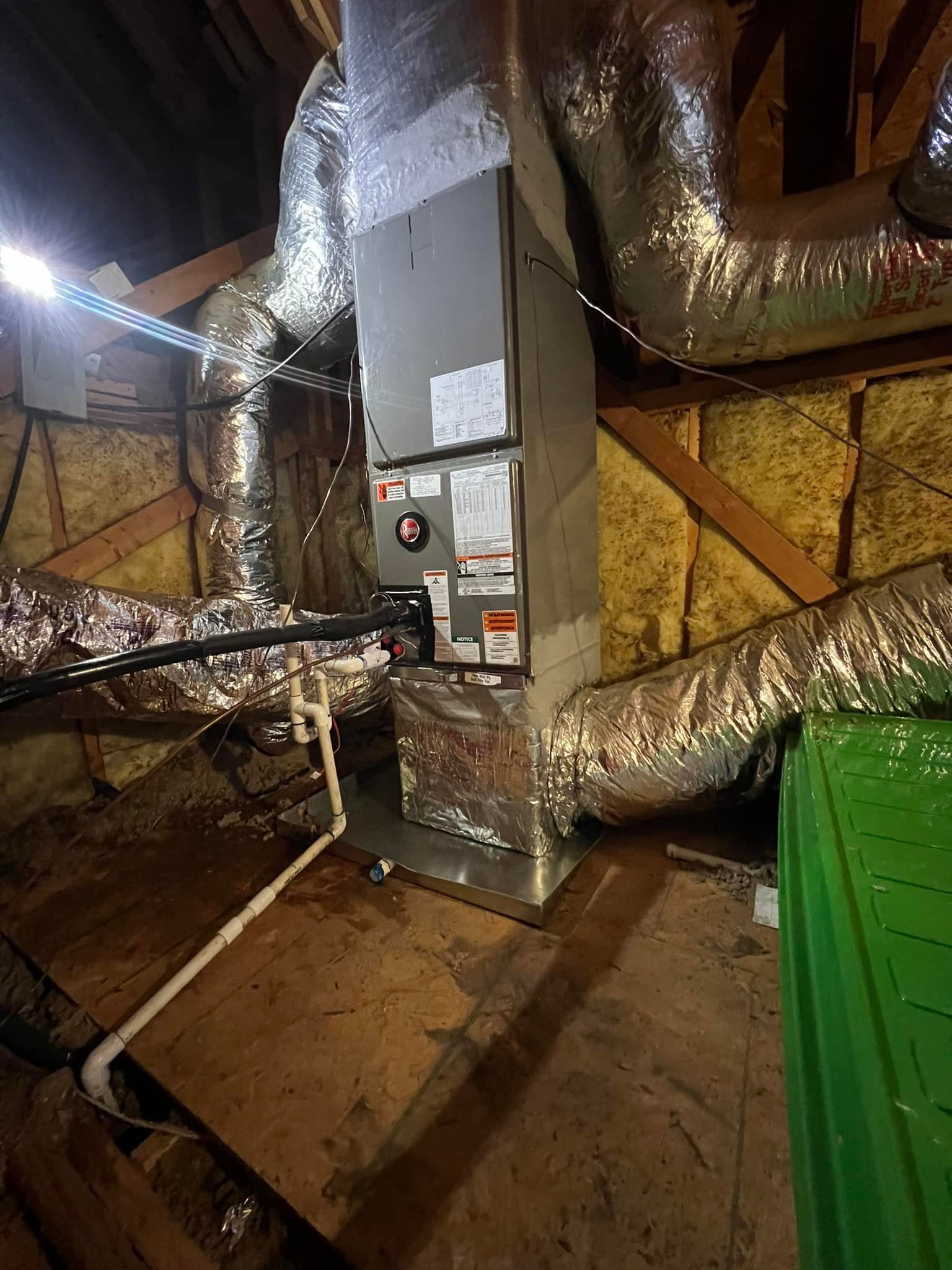 An HVAC air handler unit with silver insulation ducts installed in an attic with wooden rafters and yellow insulation.
