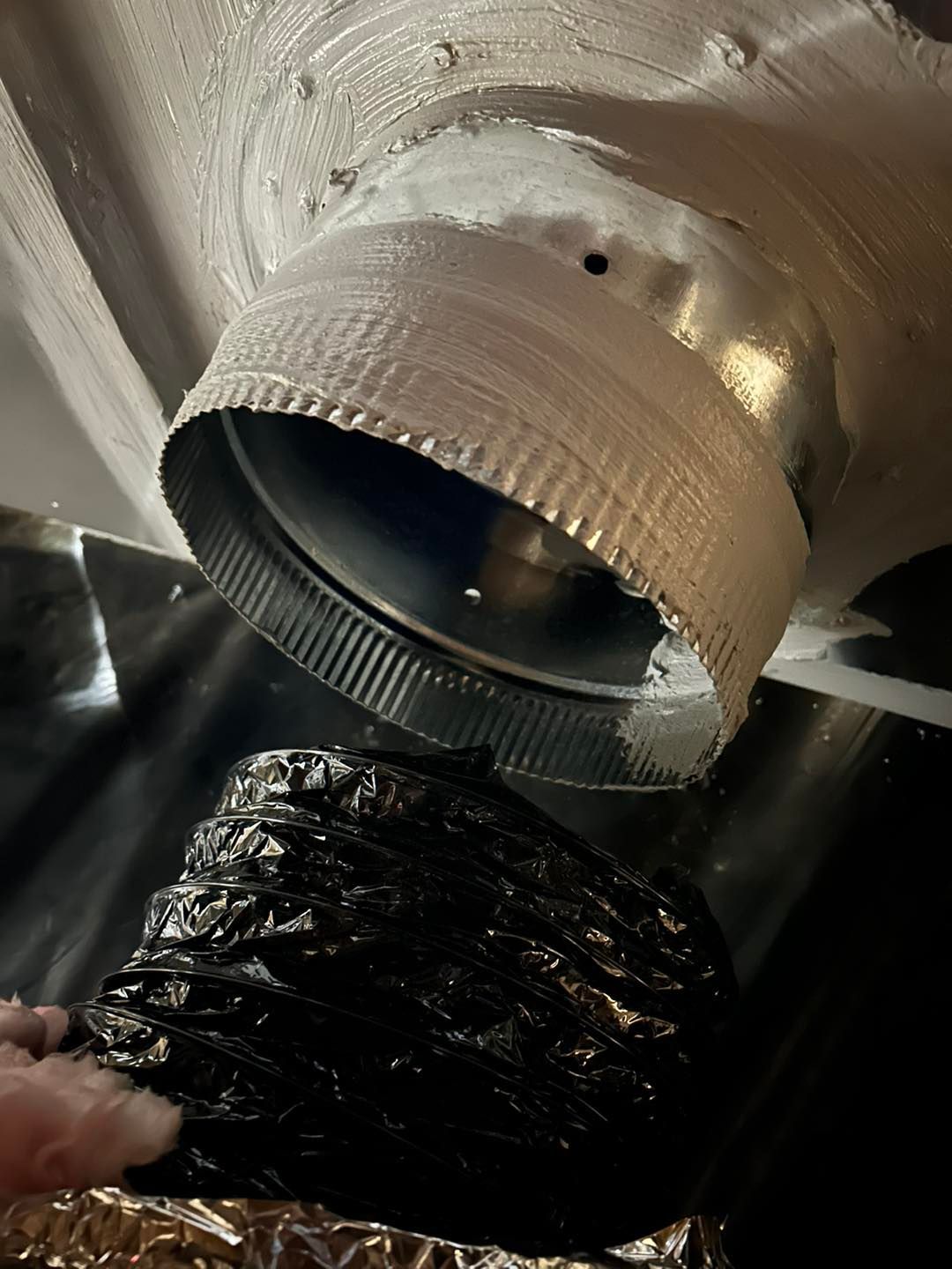 A hand holds a flexible black duct hose toward a metal exhaust port attached to a painted ceiling surface.