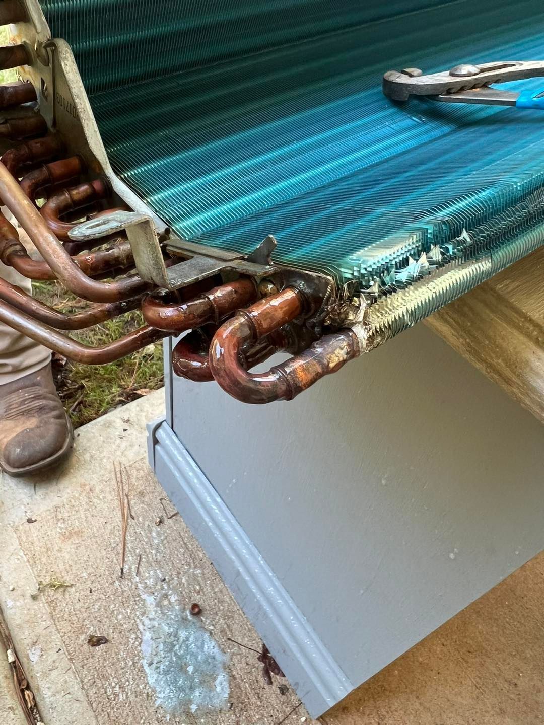 A close-up view of an HVAC copper evaporator coil with blue fins, resting on a gray cabinet outdoors on a concrete patio.