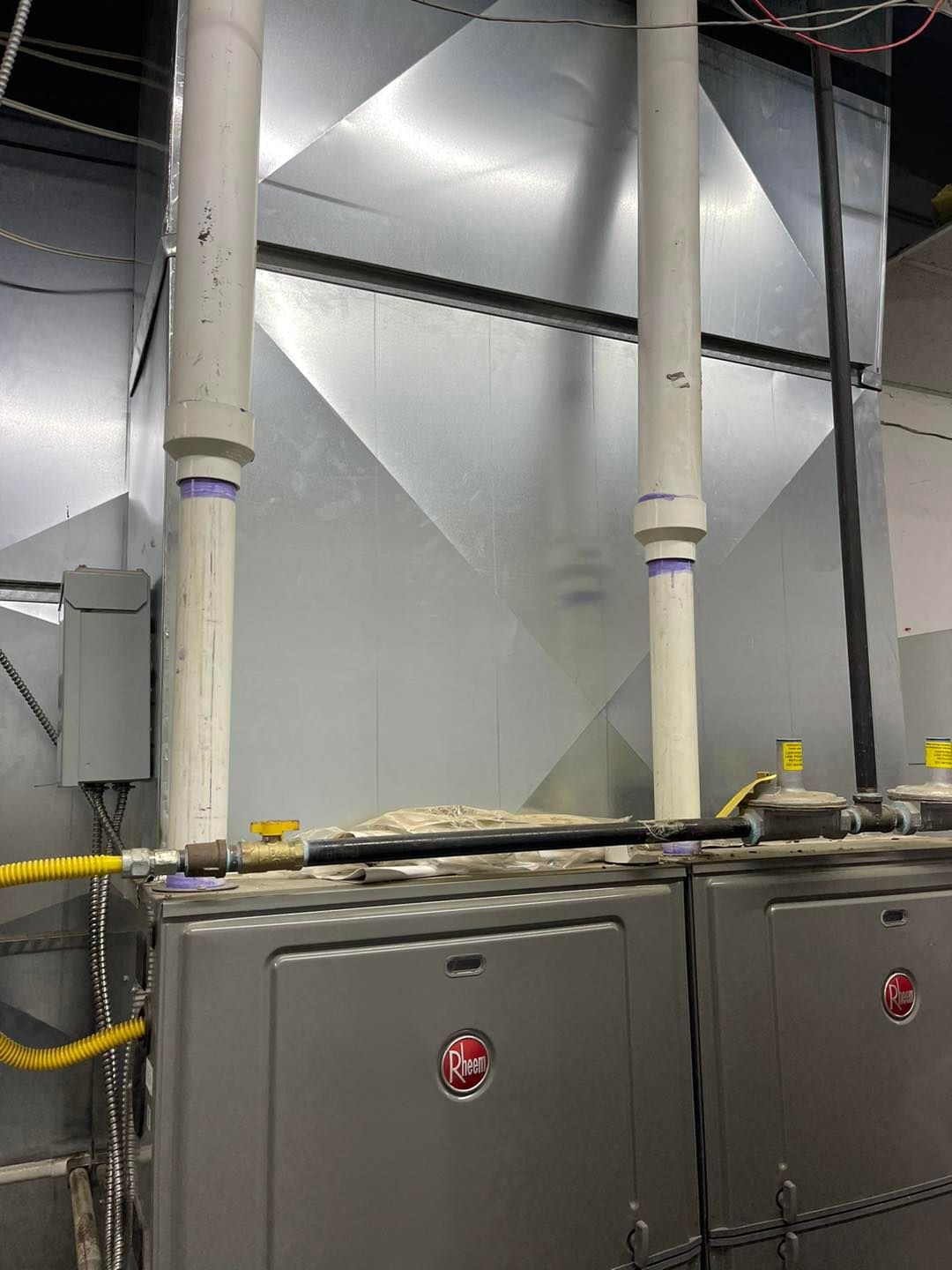 Two grey industrial HVAC furnaces with white vertical PVC exhaust pipes and a yellow gas line in a utility room.