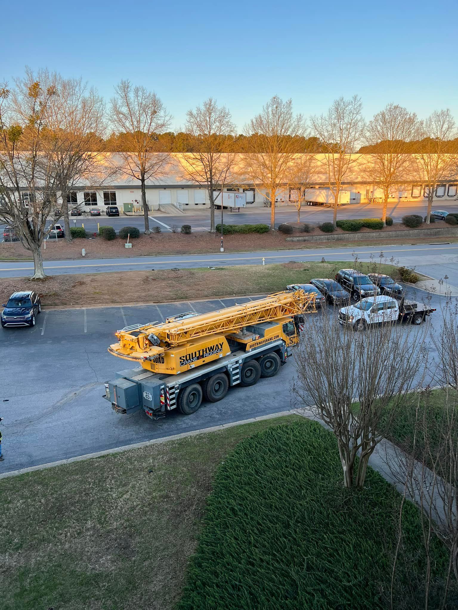 A large yellow mobile crane parked in an asphalt lot next to several passenger cars in front of a warehouse at sunrise.