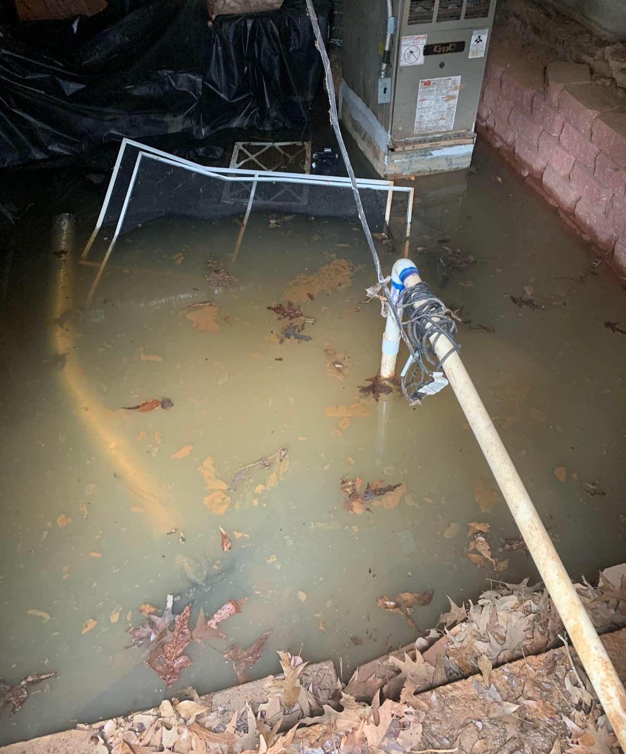 A flooded crawl space with standing brown water, an HVAC unit, and a pump hose running into the water.